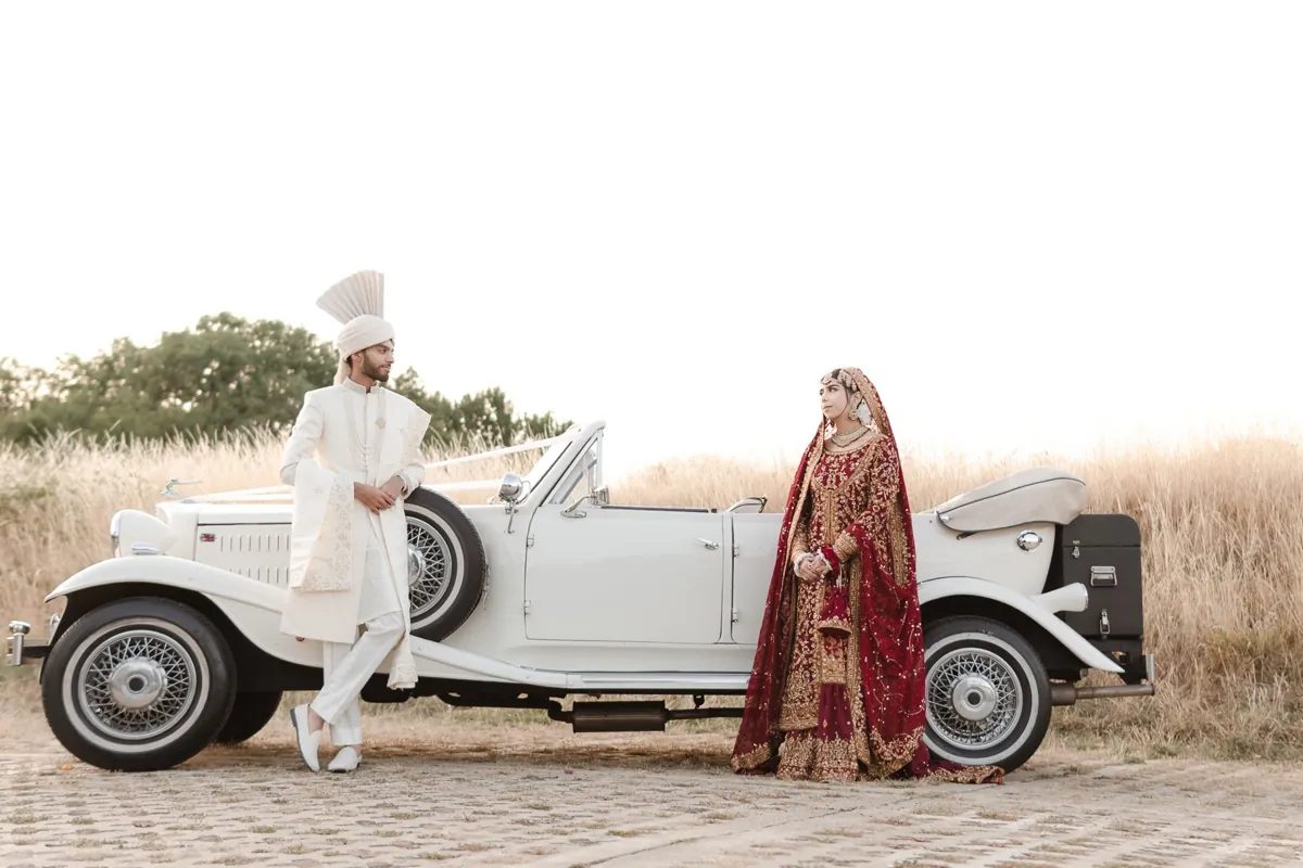 Bride and groom standing either side of a vintage wedding car in a field of tall grass