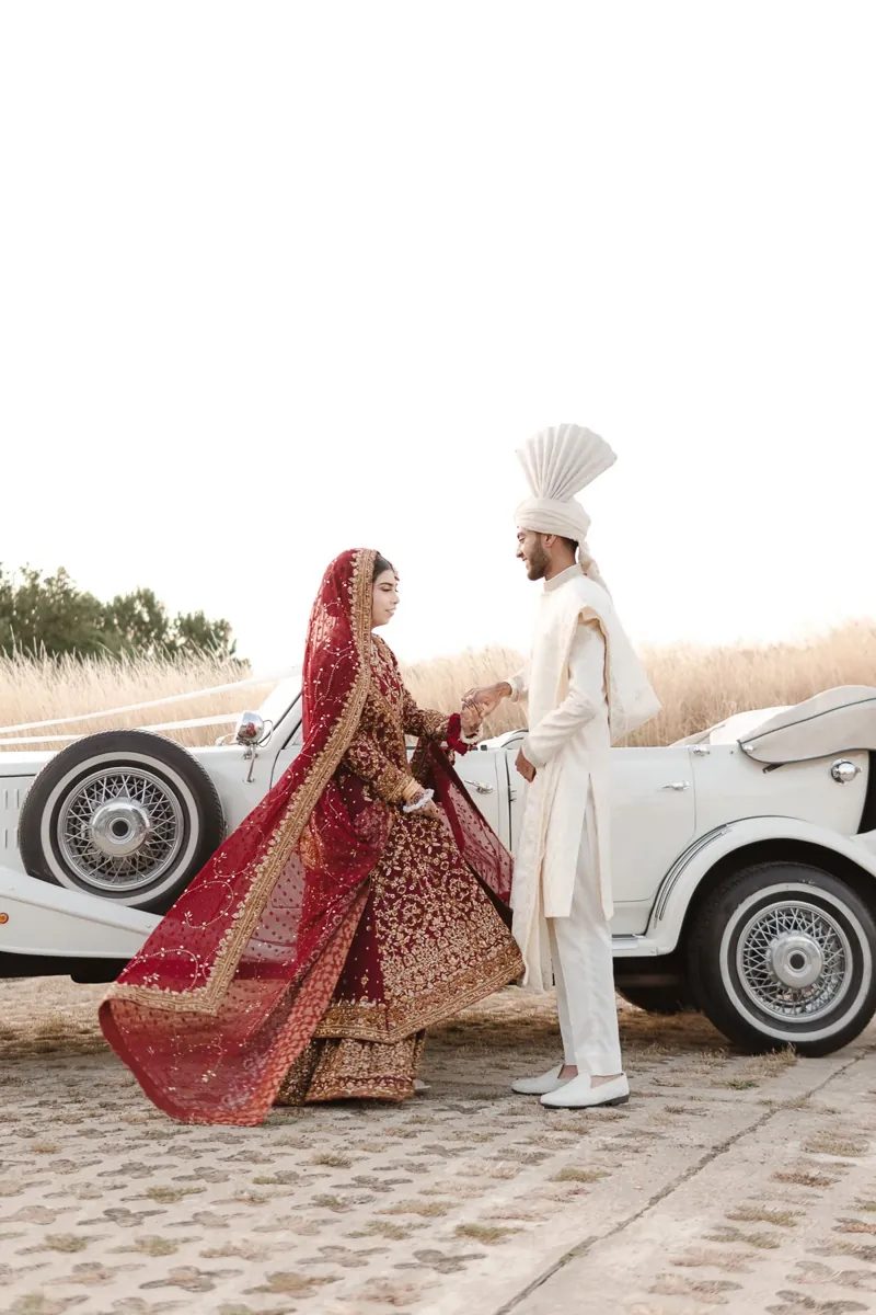 Groom holding the bride's hand beside a vintage car as her dupatta swirls