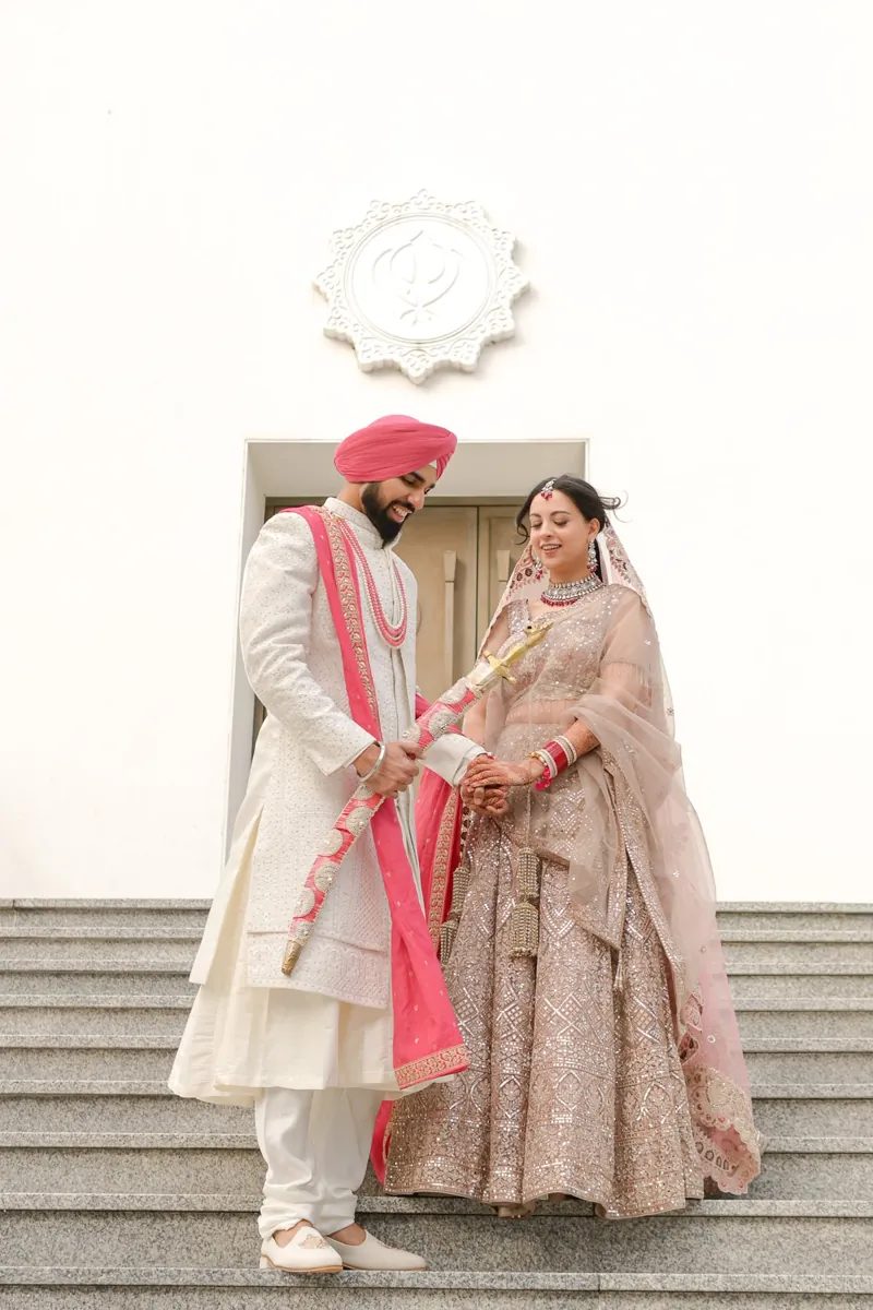 Groom in a pink turban holding a sword and laughing with the bride on the Gurdwara steps