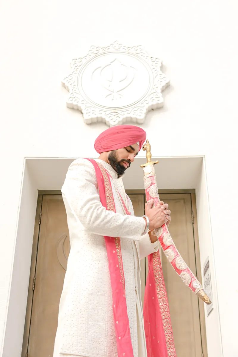 Groom in a pink turban holding a ceremonial sword outside the Gurdwara doors