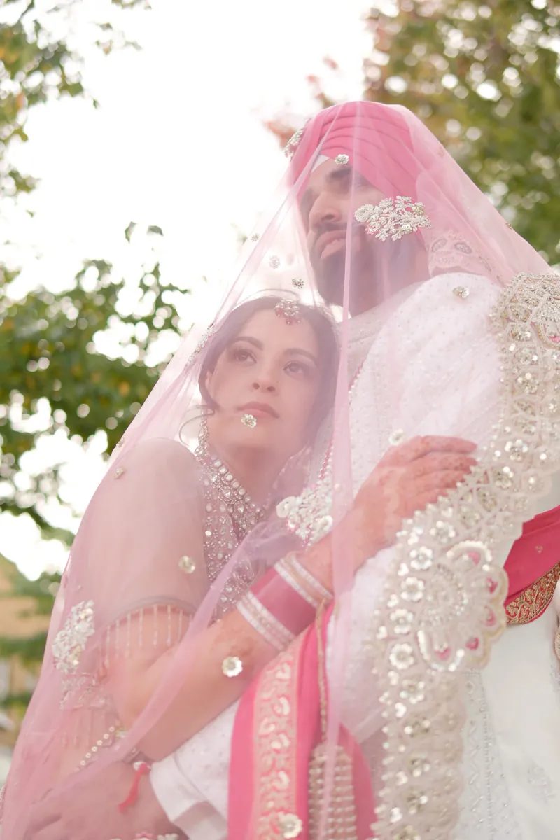 Bride resting against the groom beneath a sheer pink dupatta