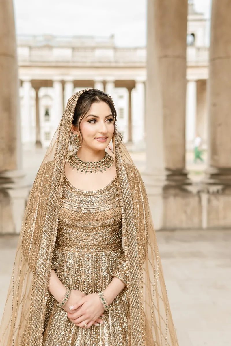 Bride in a gold lehenga and green jewellery framed by stone columns